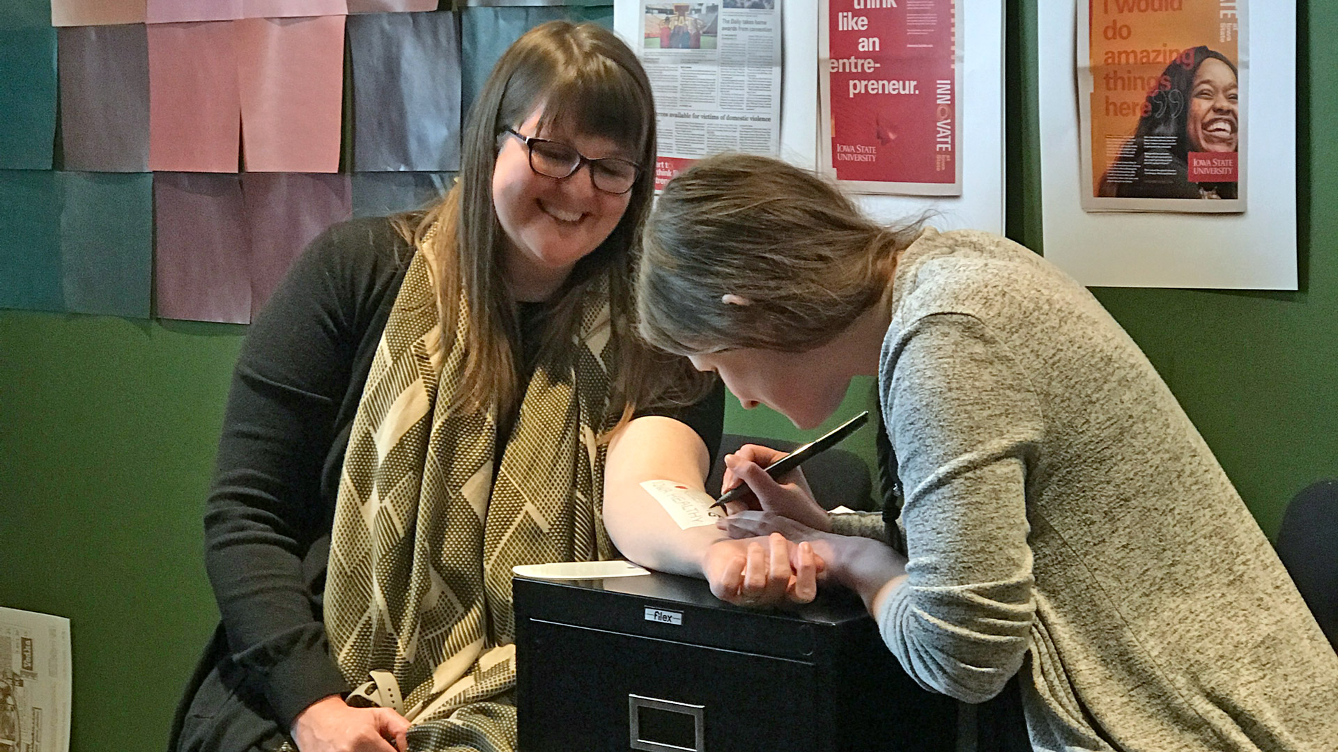Woman drawing on the inside forearm of another woman while both are smiling.